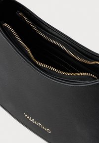 Crna kožna torba s teksturiranom površinom, dva zip odjeljka i zlatnim detaljima koji nose ugravirano ime branda "VALENTINO."