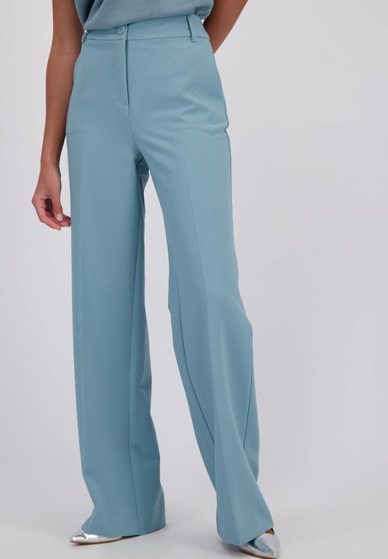 Vrouw in lichtblauwe hoge taille wijde broek met een ingestopte blauwe gebreide top en zilveren puntige schoenen.