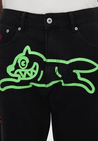 Black denim jeans featuring a large, bright green graphic of a cartoon cat with exaggerated facial features, displayed on the front.