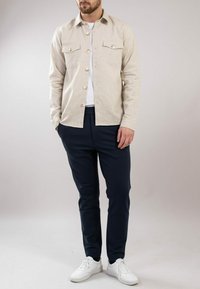 Beżowa bawełniana koszula z zapięciem na guziki i dwiema kieszeniami na piersi, zestawiona z granatowymi spodniami slim-fit i białymi sneakersami, na neutralnym tle.