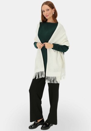 Biały szal z frędzlami zarzucony na ciemnozielony sweter, z fakturowanym wzorem wzdłuż krawędzi; noszony z czarnymi spodniami o szerokich nogawkach i błyszczącymi butami.