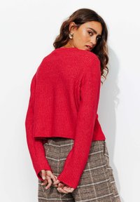 Pull rouge côtelé avec des manches longues, une taille cropped et un tissu texturé. Associé à un pantalon à carreaux marron. Fond clair.