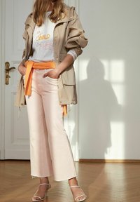 Beżowa lekka kurtka, szara koszula z pomarańczowym napisem, różowe rozszerzane spodnie, pomarańczowy sweter związany w talii i białe sandały na paski.