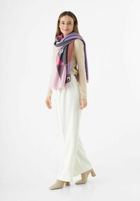 Écharpe multicolore avec des motifs abstraits dans les tons rose, violet et marron, fabriquée en tissu doux, drapée sur un pull beige clair et accompagnée de pantalons crème.