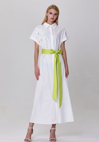 Robe blanche à manches courtes, avec un détail textur�é floral sur un côté et une ceinture vert vif nouée à la taille. Tissu lisse.