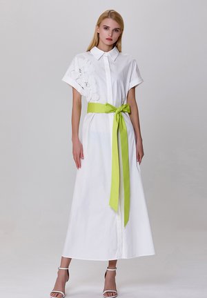 Robe blanche à manches courtes, avec un détail texturé floral sur un côté et une ceinture vert vif nouée à la taille. Tissu lisse.