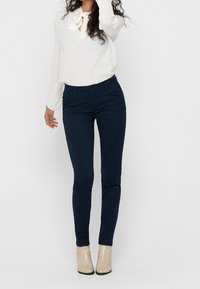 Granatowe spodnie slim fit z bawełny o gładkiej strukturze; zestawione z lekką białą bluzką i beżowymi szpilkami.