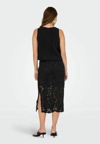 Schwarzes ärmelloses Top mit einem Reißverschluss, kombiniert mit einem langen schwarzen Spitzenrock mit floralen Mustern und seitlichen Schlitzen. Einfache schwarze Absätze.