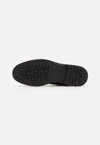 Semelle d'une botte noire avec des motifs de crampons profonds comprenant des croix et des lignes pour l'adhérence, présentée sur un fond blanc uni.