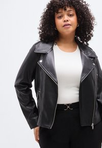 Μαύρο μπουφάν μηχανής από συνθετικό δέρμα με φερμουάρ, φαρδύ γιακά και πλαϊνές τσέπες, φορεμένο πάνω από ένα λευκό ριμπ μπλουζάκι και μαύρη ζώνη.