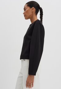 Giacca nera con zip, dalla texture liscia, colletto rotondo e maniche ampie, abbinata a pantaloni chiari.