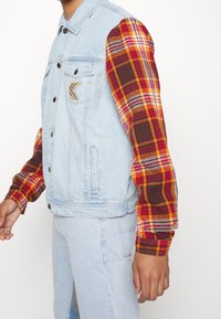 Denim vest with plaid long sleeves in red, orange, and brown. Features a small logo on the chest and metallic buttons. Light wash color.