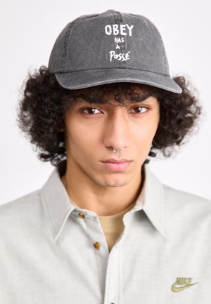 Szürke baseball sapka, fehér hímzéssel az "OBEY HAS A POSSE" felirattal; világos pólóval viselve, amelyen egy kis zöld Nike logó található.
