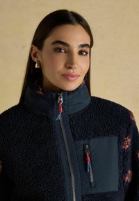 Marineblauwe fleecejas met gestructureerde stof, hoge kraag en zakken met rits. Voorzien van rood en blauw ritstrekkers, bloemaccenten op de mouwen.