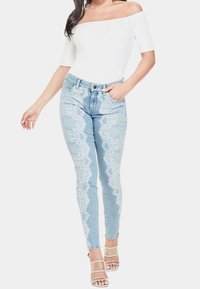 Jeans de mezclilla azul claro con un patrón de encaje y una cintura alta, que presentan un ajuste slim. Combinados con una blusa blanca sin hombros y tacones con tiras.