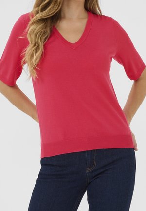 Nainen, jolla on pitkät vaaleat hiukset, pukeutunut kirkkaan vaaleanpunaiseen lyhythihaiseen V-kaulaiseen neuleeseen ja tummansinisiin farkkuihin, kädet levitettyinä lanteille.