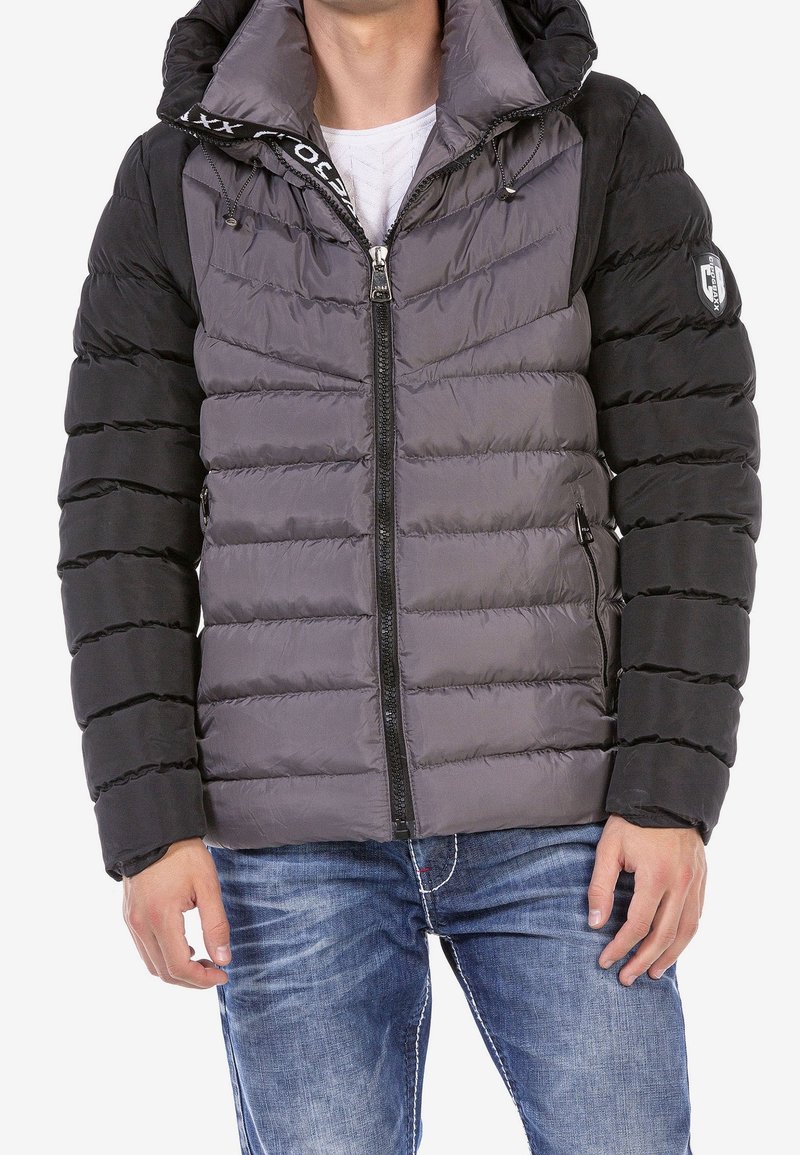 Chaqueta acolchada en gris y negro, con cierre de cremallera en el frente, capucha y puños elásticos. Superficie texturizada con costuras horizontales y bolsillos laterales.