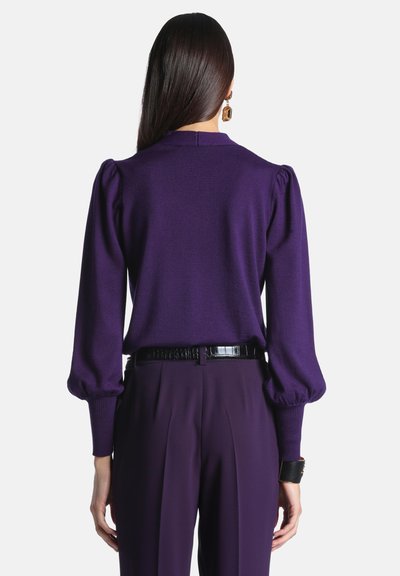 Femme vue de dos portant un haut violet à manches longues avec des épaules bouffantes et un pantalon violet assorti avec une ceinture noire.