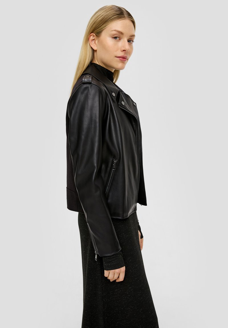 Schwarze Leder-Motorradjacke mit tailliertem Design, silbernen Beschlägen, Reißverschlüssen und Schulterklappen, kombiniert mit einem dunklen Langarm-Oberteil.