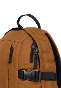 Zaino in tessuto marrone con cerniere e fibbie nere. Presenta una maniglia superiore e una texture data dalle pannellature cucite lungo i lati.