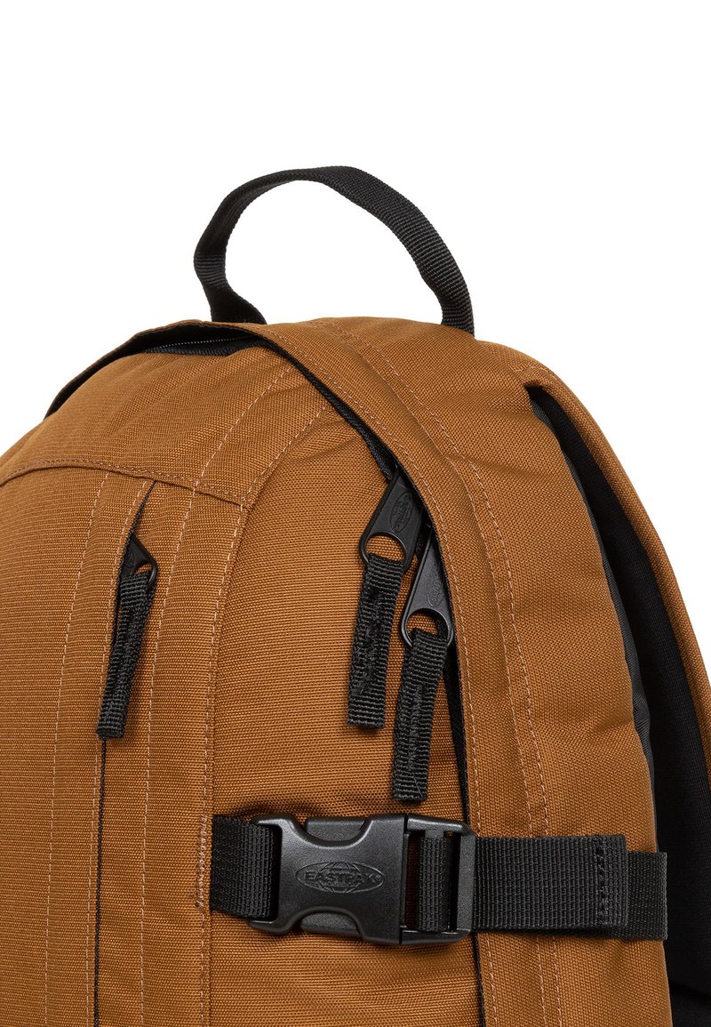 Sac à dos en tissu marron avec des zips et une boucle noirs. Il possède une poignée supérieure et une texture provenant de panneaux cousus le long des côtés.
