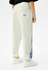 Osoba v bílých teplácích se šedým bočním panelem a černými knoflíky, s elastickými manžetami, zadní kapsou s etiketou, v kombinaci s bílými teniskami.