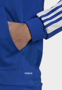 Sudadera deportiva azul con un bolsillo frontal, puños acanalados y acentos de tres rayas blancas en las mangas. Presenta la marca AEROREADY en el dobladillo.