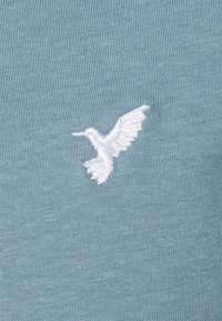 Light blue knit fabric featuring a white embroidered hummingbird design, positioned slightly off-center.