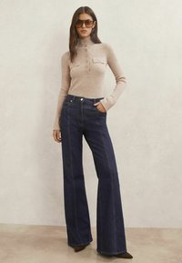 Bézs bordás pulóver magas nyakkal, gombos részletekkel és foltzsebekkel, sötétkék, kontrasztos varrással díszített bővülő nadrággal.