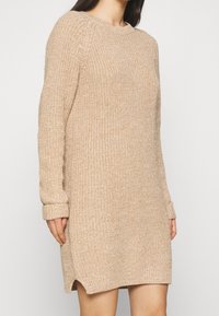 Robe-pull beige en maille avec manches longues, poignets côtelés, fentes latérales et coupe décontractée. Le tissu texturé ajoute un intérêt visuel.