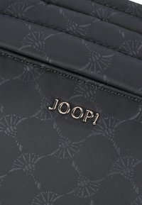 Borsa in tessuto nera con un motivo sottile. Presenta un logo metallico dorato "JOOP!" sul davanti. Superficie testurizzata e bordi cuciti.
