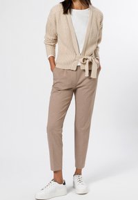 Knit beige cardigan with a tied waist, paired with light brown tapered pants and white sneakers with black accents. Simple, textured design.