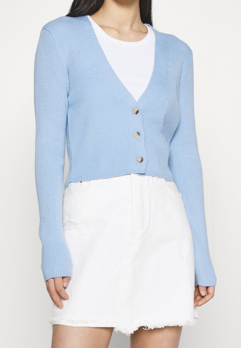 Femme portant un cardigan court bleu clair boutonné par-dessus un haut blanc et une jupe en jean blanche à ourlet effiloché.