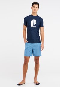 Námořnický rash guard s bílým logem "PROTEST", zkombinovaný se světle modrými plavkami. Žabky dokončují uvolněný plážový vzhled.