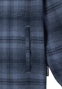Camisa a cuadros azules con bolsillo en el pecho, diseño de botón y puños acanalados. Etiqueta visible en el costado con el nombre de la marca.
