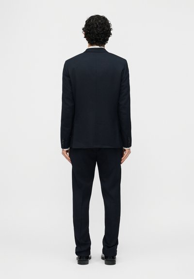 Άνδρας με σγουρά μαλλιά στέκεται γυρισμένος με την πλάτη φοράει εφαρμοστό σκούρο μπλε κοστούμι με μαύρα παπούτσια σε απλό ανοιχτό φόντο.