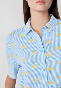 Blauw en wit gestreepte kortebloezende shirt met een gele fruitprint, kraag en knoopsluiting. Zachte stoftextuur.