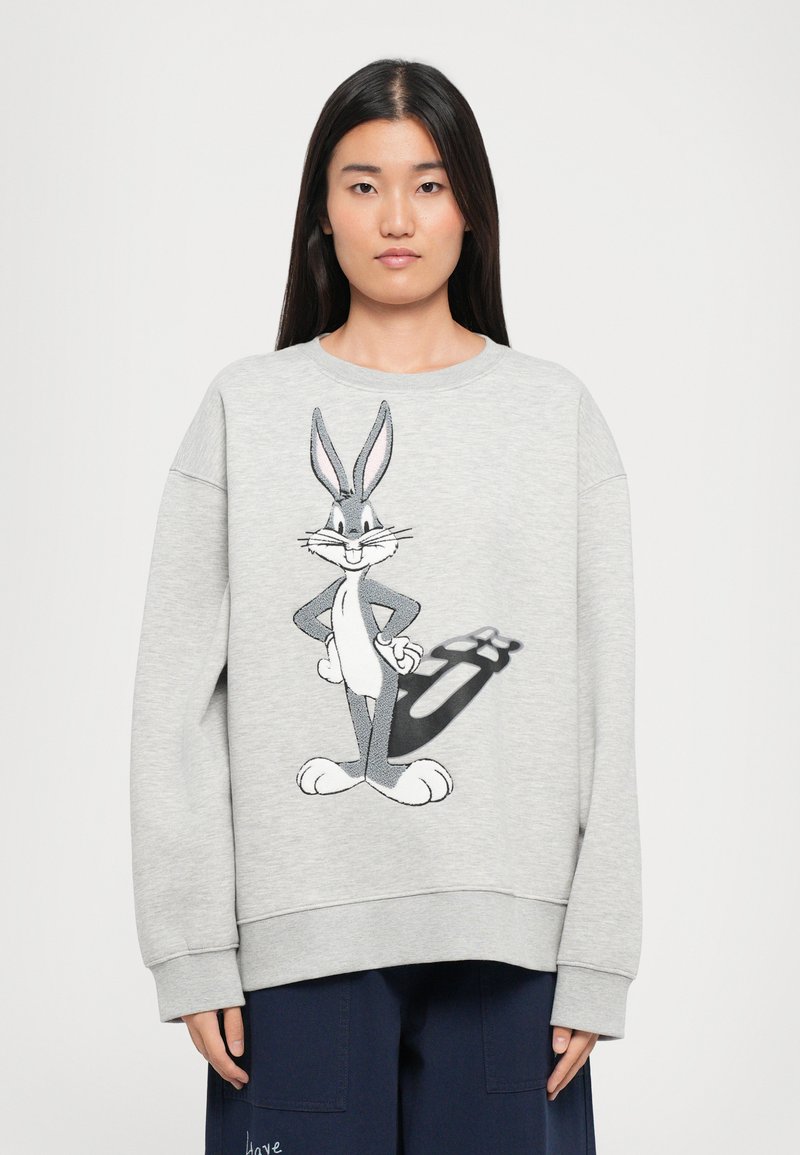 Žena v sivom mikinovom svetre s veľkým grafikou Bugs Bunnyho, stojí na pozadí s jednoduchým bielym pozadím.
