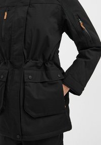 Schwarze Outdoor-Jacke mit tailliertem Schnitt, mehreren Taschen, Reißverschluss und Druckknopfverschlüssen, aus einem glatten, strukturierten Stoff.