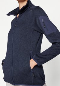 Chaqueta de forro polar azul marino con cuello alto, cierre frontal con cremallera, bolsillos laterales y bolsillo en la manga con cremallera. Tejido texturizado y diseño ajustado.