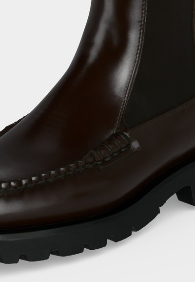 Gros plan d'une botte en cuir marron foncé brillant avec des détails cousus et une semelle épaisse en caoutchouc noir à crampons profonds.