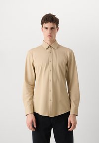 Camisa de botones de manga larga beige con tela texturizada, cuello puntiagudo y seis botones en la parte delantera. Combinada con pantalones oscuros.