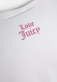 Balta medvilninė marškinėliai trumpomis rankovėmis su rožinės spalvos „Love Juicy“ užrašu stilizuotu šriftu arti iškirpės. Minkšta tekstūra.