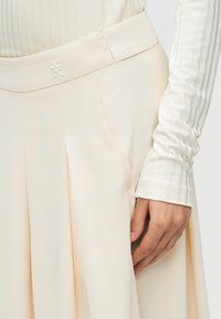 Μέση και χέρι ατόμου που φοράει κρεμ πλισέ φούστα και λευκό ριμπ πλεκτό τοπ με μακριά μανίκια, εμφανίζοντας πλευρικό φερμουάρ και μικρό λογότυπο στη μέση της φούστας.