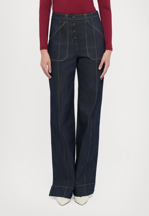 Brede, donkere spijkerbroek met een hoge taille, voorzien van gouden stiksels en een knoopsluiting. Gecombineerd met puntige witte schoenen.