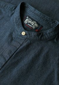 Donkerblauwe jeanshemd met een mandarinokraag, voorzien van een enkele knoopsluiting en een label met de tekst "SuperDry, gemaakt in India."