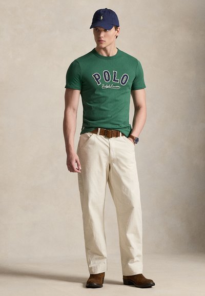 Πράσινο βαμβακερό μπλουζάκι με λογότυπο "POLO", κρεμ chinos, καφέ ζώνη και καφέ υποδήματα, συνοδευόμενο από μπλε σκούφο.