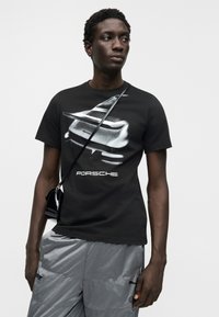 Μαύρο μπλουζάκι με γκρι γραφικό αυτοκινήτου Porsche και λευκό κείμενο. Το μοντέλο φορά γκρι παντελόνι και κρατά μια μικρή μαύρη τσάντα χιαστί.