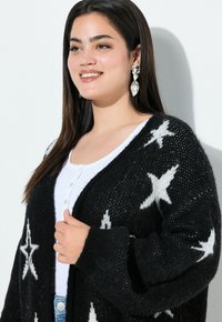 Cardigan noir en tricot avec des motifs d'étoiles blanches, à ouverture frontale, manches longues, tissu texturé, associé à un haut blanc.