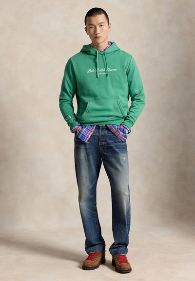Άνδρας με πράσινο φούτερ Polo Ralph Lauren, καρό πουκάμισο, ξεβαμμένο μπλε τζιν και καφέ μπότες με κόκκινα κορδόνια, στέκεται με τα χέρια στις τσέπες.